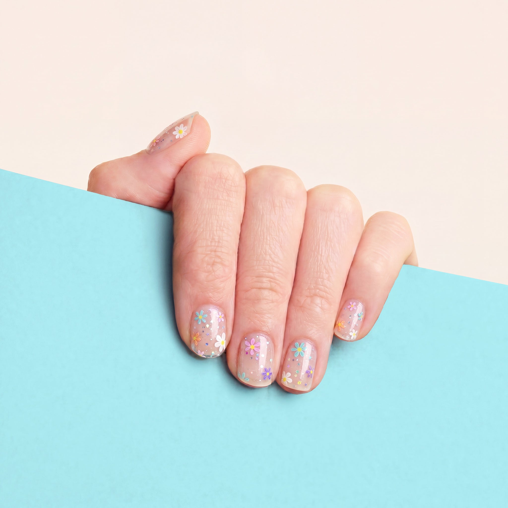 Hand with clear mesme gel nail wraps featuring colourful daisy flowers against a blue background.