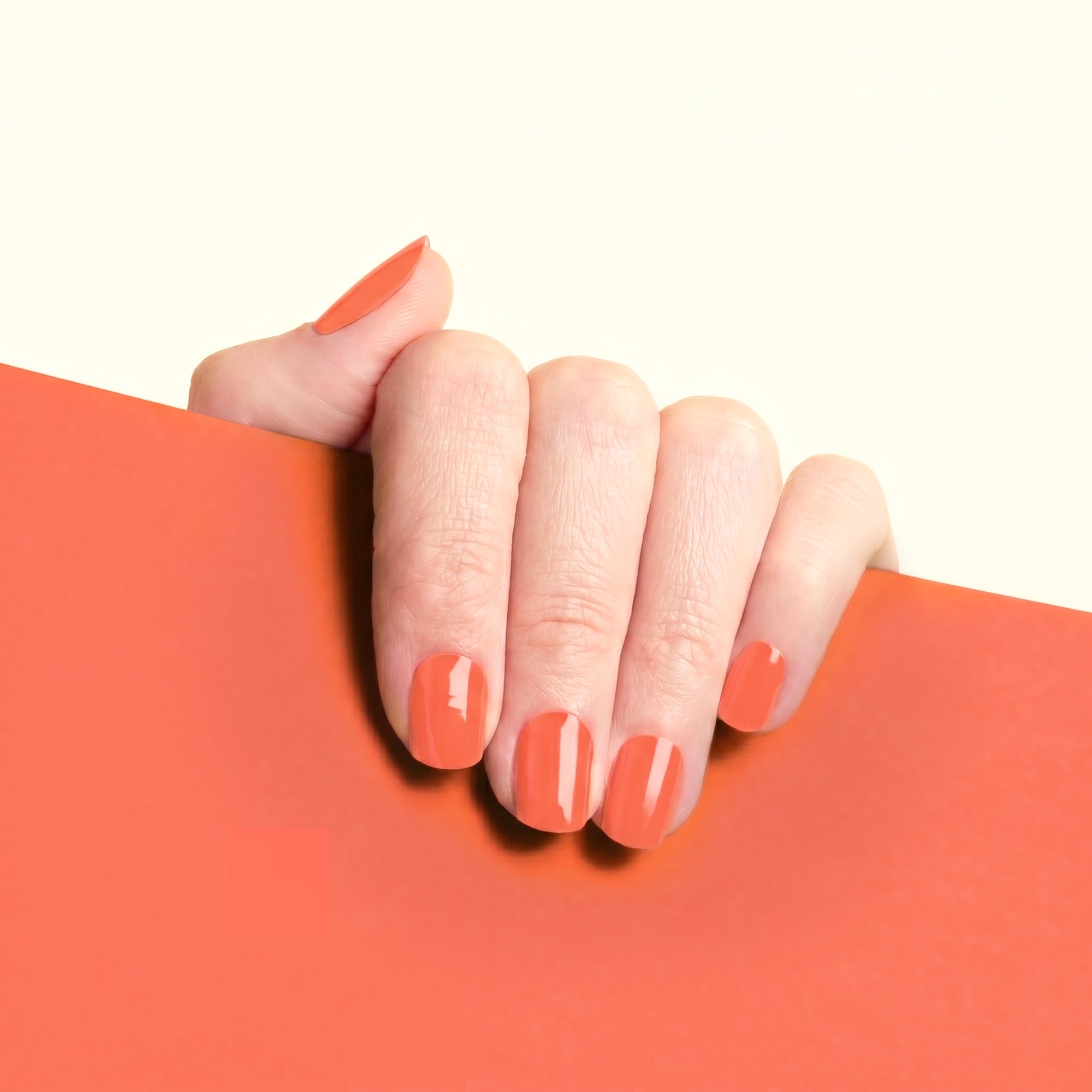 Hand with glossy coral-red Suzanne mesme gel nail wraps against a matching warm red background.