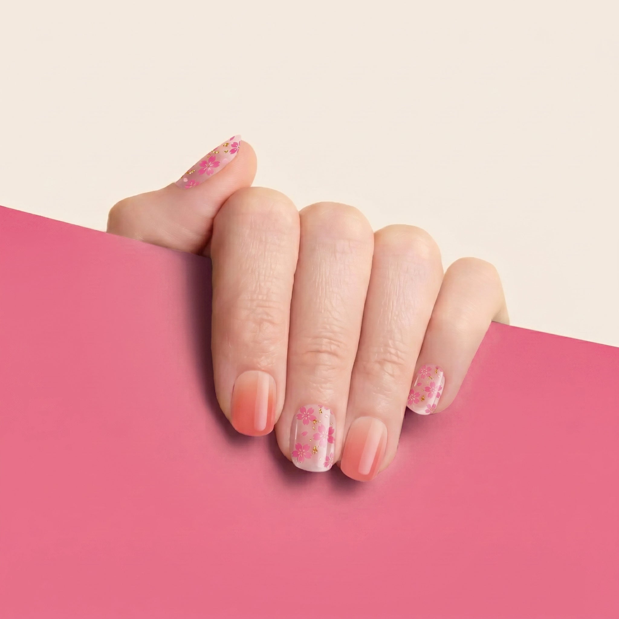 Hand with peach-pink ombré mesme gel nail wraps with pink flowers and gold accents against a pink background.
