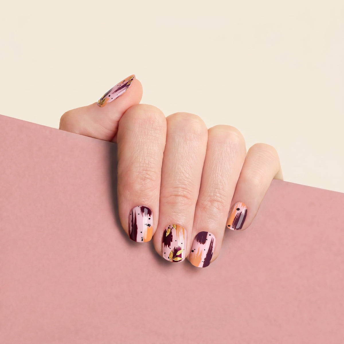 A close-up of a hand with mesme nail wrap in a modern abstract design featuring dark plum, peach, orange, and gold splatter and stripe patterns, resting on a dusty pink surface against a light cream background.